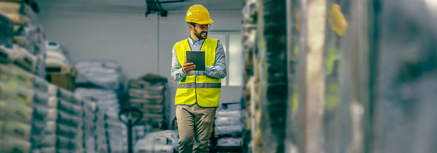 warehouse supervisor in vest and protective helmet walking around warehouse and using tablet.